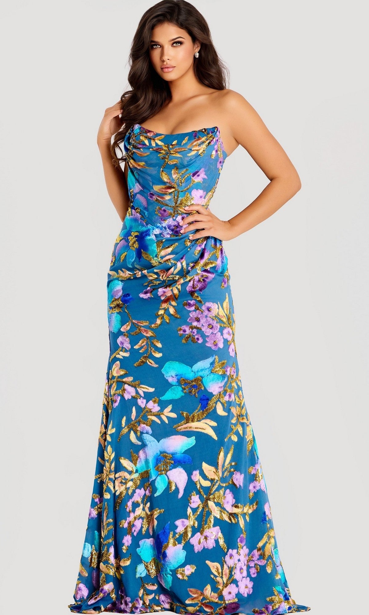 A woman stands against a plain light background in the Jovani 47654, a long blue strapless prom dress with a fitted bodice, slight draping detail, and floor-length fabric adorned with delicate floral patterns.